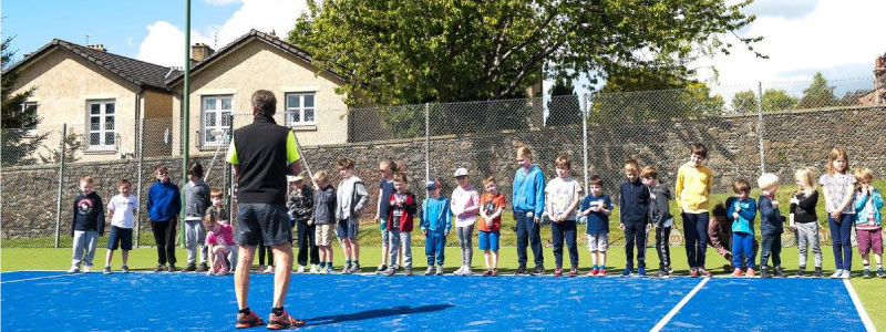 Children lined up on an outdoor tennis court with a coach standing in front of them
