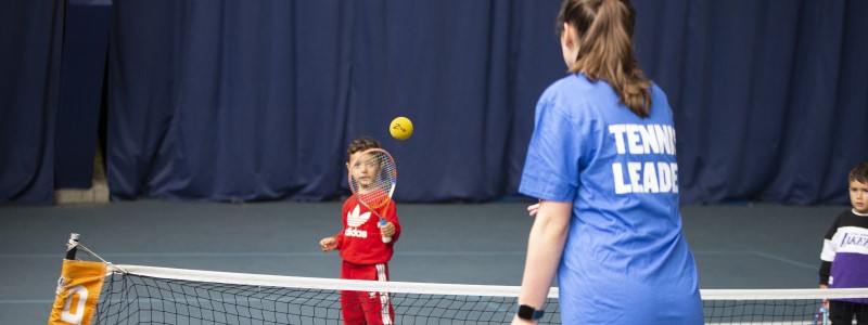 A girl wearing a blue t-shirt with Team Leader printed on the back throws a foam ball to a young boy wearing red and holding a tennis racket